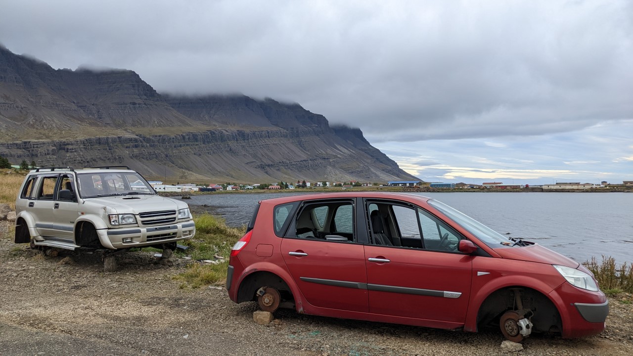 Ein silbernes und ein rotes Fahrzeug Wrack ohne Reifen in Island. Im Hintergrund ist eine Bucht und hohe, dunkle, in den Wolken verschwindende Berge.  