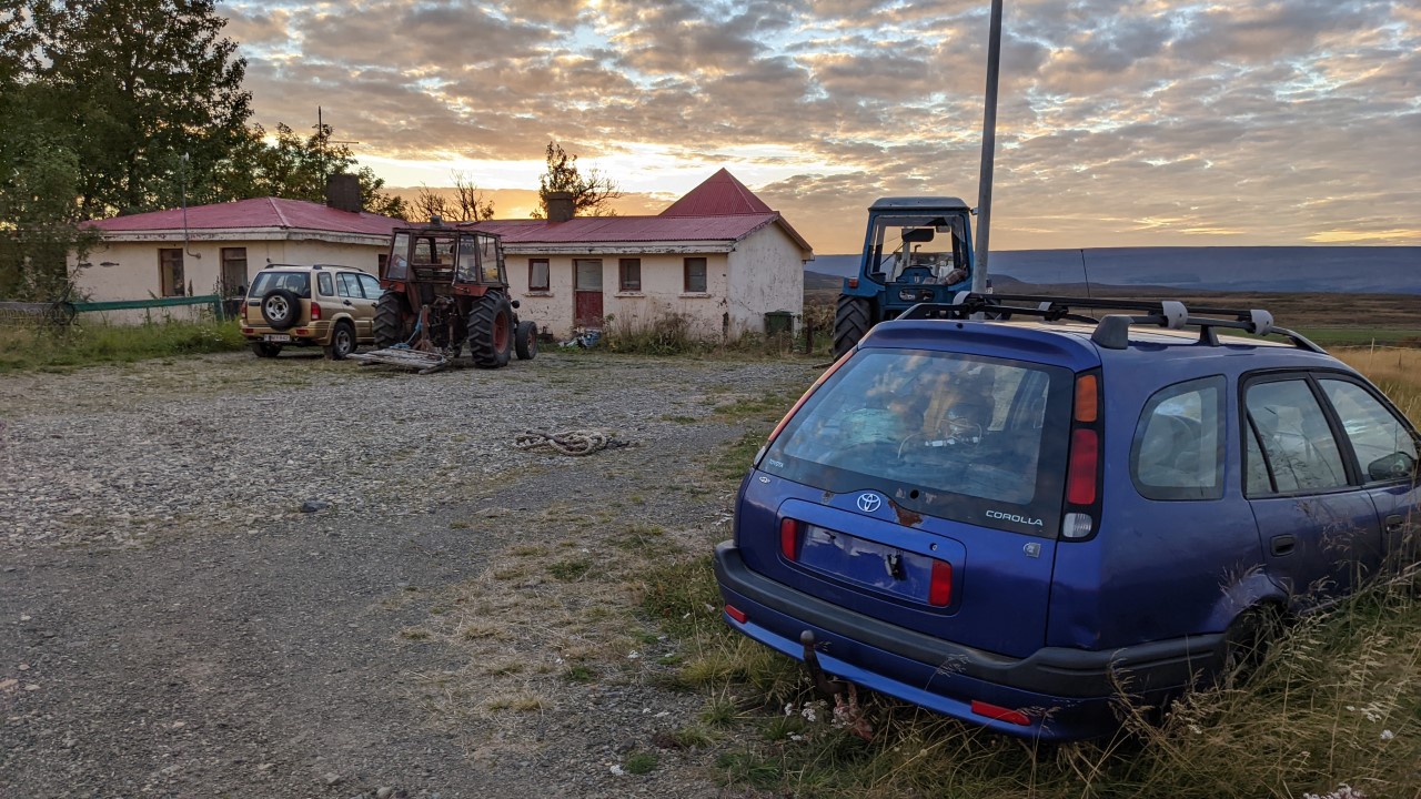 Zwei alte Trecker und ein blauer Toyota Corolla ohne Nummernschild parken vor einem leer stehenden Haus.  
