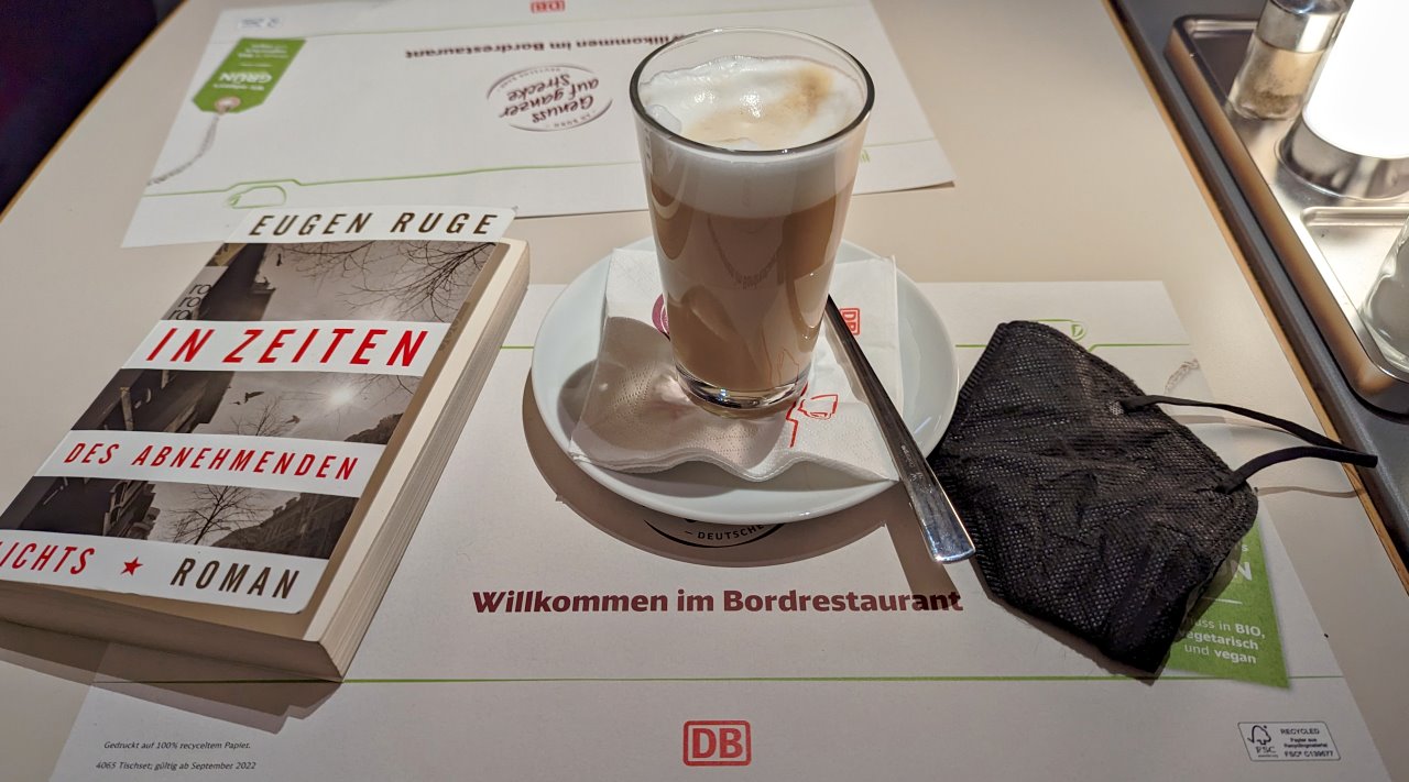Das Buch "In Zeiten des abnehmenden Lichts" neben einer Corona Masken und einen Milchkaffee fotografiert in einem ICE Zugrestaurant