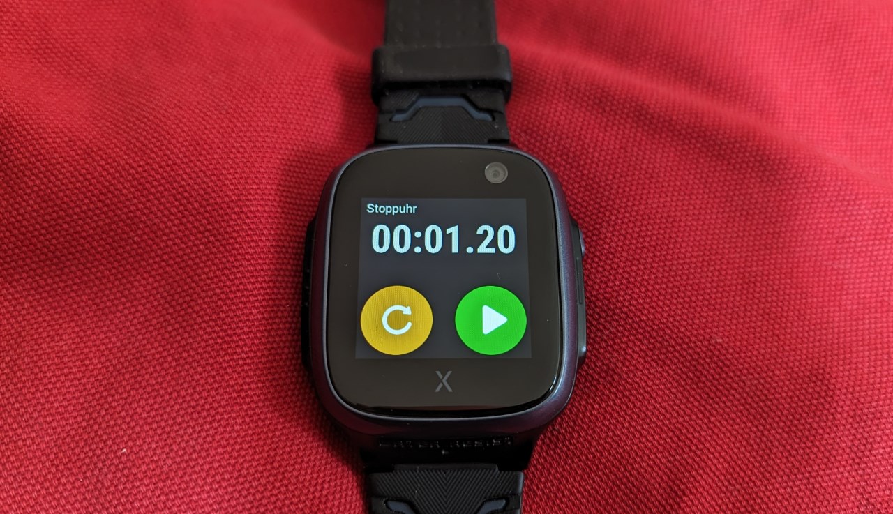 Xplora x5 Smartwatch auf einem roten Kissen, der Bildschirm zeigt die Stopuhr mit orangenem Reset Button und grünem Start Button.  