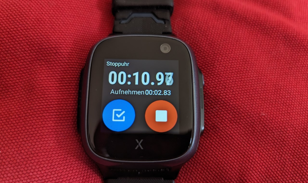 Xplora x5 Smartwatch auf einem roten Kissen. Der Bildschirm zeigt die Stoppuhr mit einem blauen Zwischenzähler Button und rotem Stop Button. 