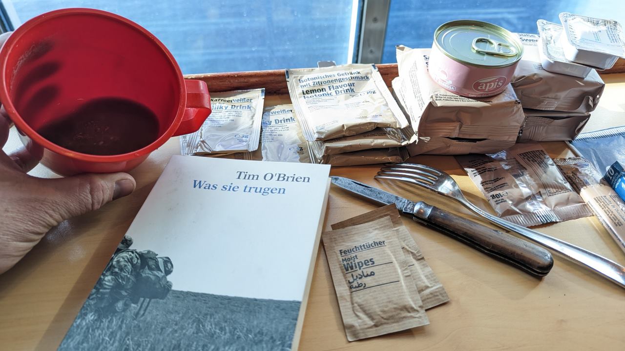 Ein roter Plastikbecher mit einem Rest Kaffee, das Buch Tim O'Brien was sie trugen, Bundeswehr EPA Nahrungsrationen, Messer und eine Gabel. 