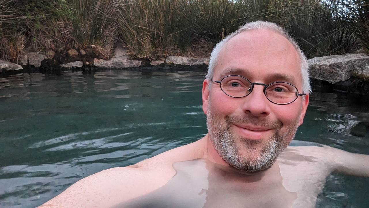 Ein zufrieden aussehender männlicher Abenteurer mit Dreitagebart und Brille sitzt in einem natur Badebecken mit 40 Grad heissem Wasser  