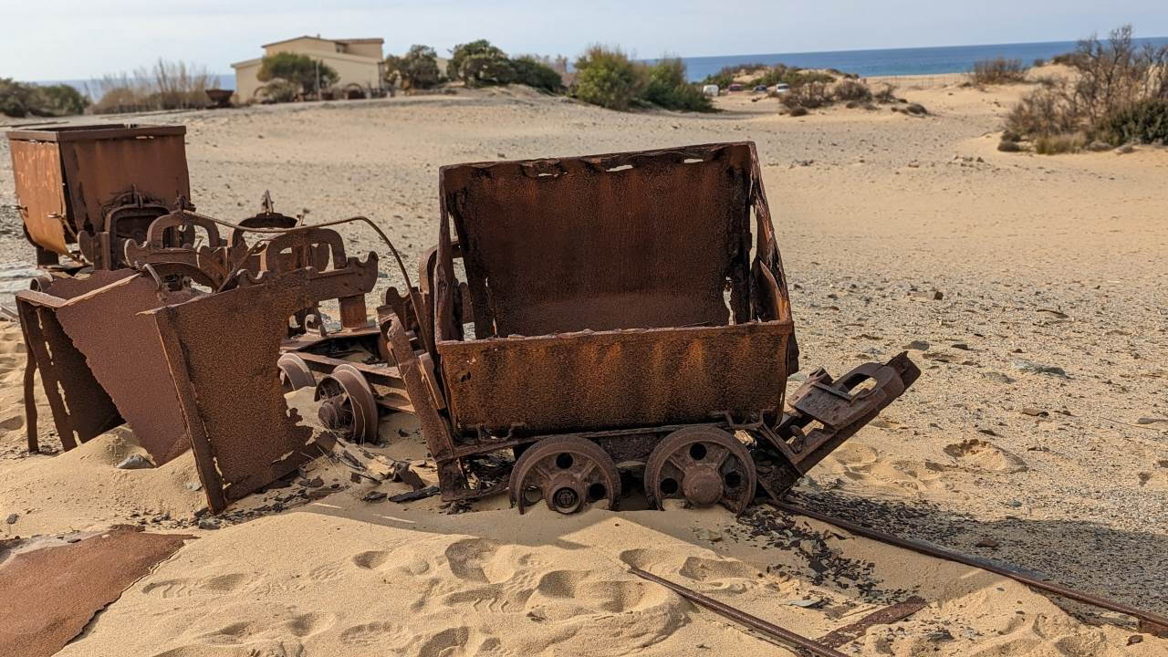 Eine entgleiste Lore steht quer zu im Sand versunkenen Eisenschienen in Le Dune la Piscinas Sardinien