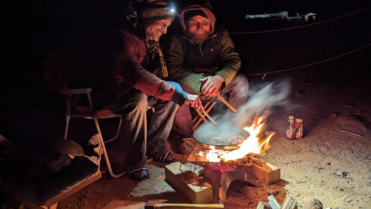 Zwei Freunde sitzen Nachts mit zufriedenem Gesichtsausdruck gemeinsam an einem Holz Lagerfeuer, auf dem in einer Pfanne etwas brutzelt. 