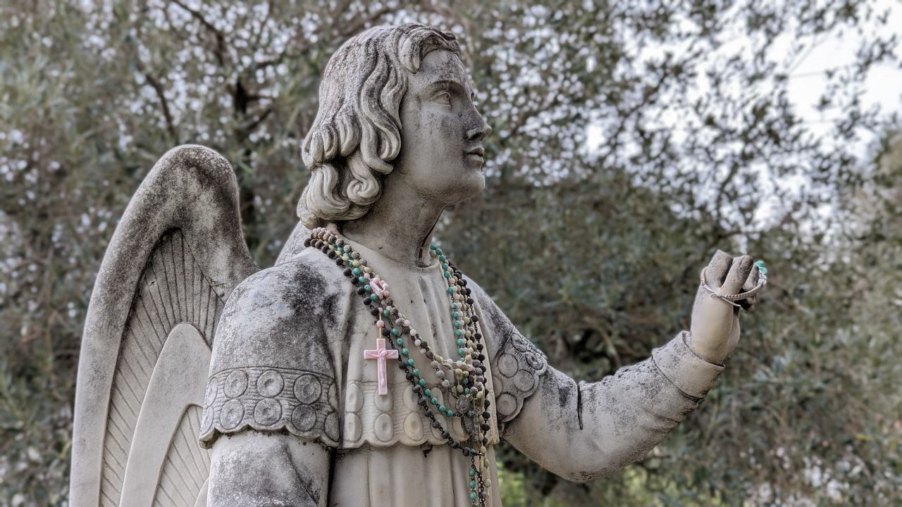 Eine weisse männliche Engels Statue mit Flügeln geschmückt mit einer grünen Perlen Kette und einem rosa Kreuz