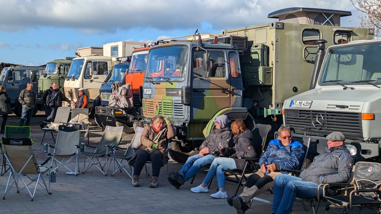 Eine Gruppe Menschen sitzt in Gartenstühlen im Sonnenschein vor etwa sieben LKW