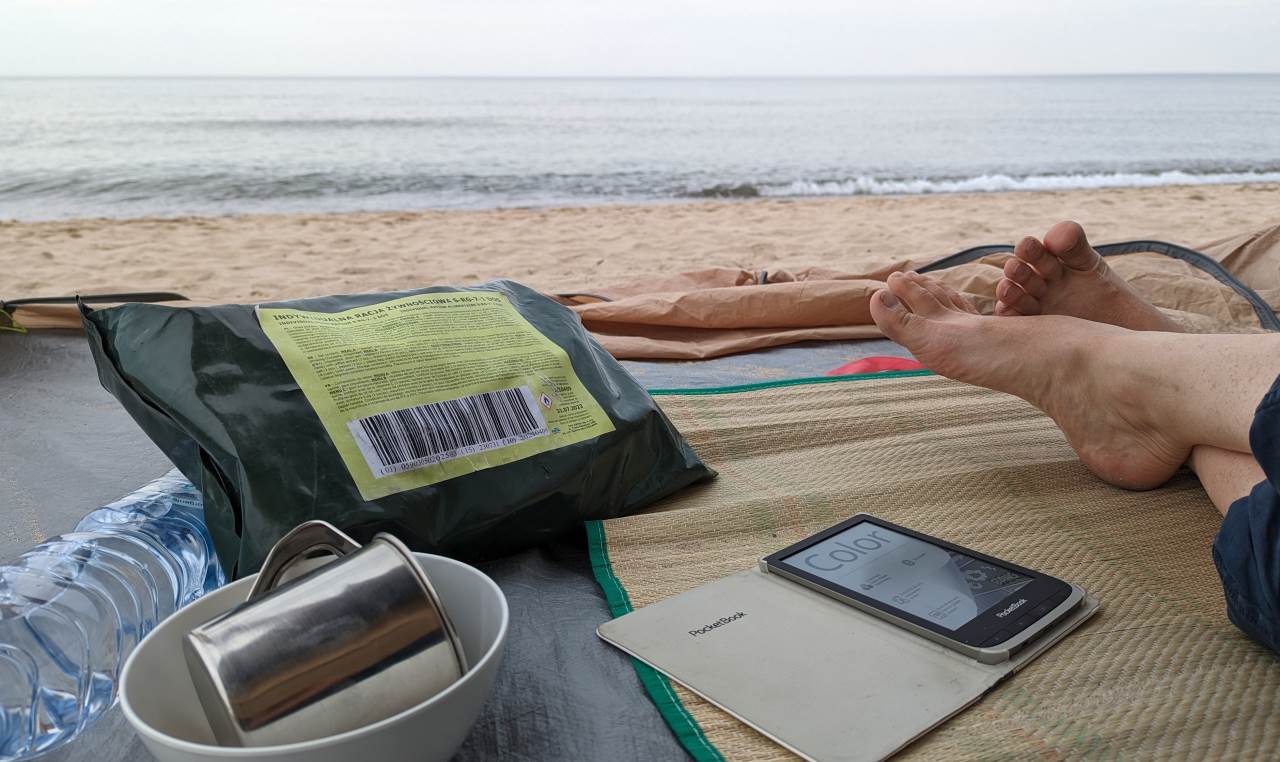 Blick aus der Strandmuschel auf dem Ostsee Strand in Polen, bedeckter Himmel. Im Vordergrund liegen ein eBook Readder, ein 24 EPA und ein silberner Becher in einer weissen Schale. 