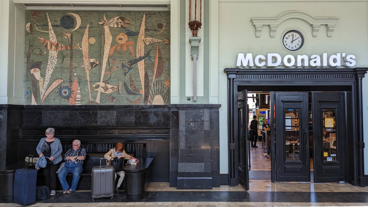 Im Bahnhof. Drei Menschen sitzen vor einem McDonalds Eingang unter einem alten Mosaik Bild