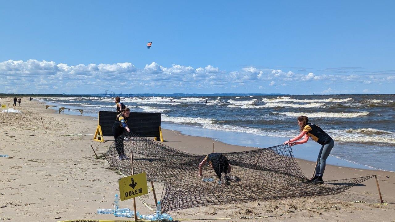 Eine Fitness Übung am Strand von Sobieszwska in Polen. Teilnehmer klettern am Strand unter einem schwarzen Netz hindurch und überklettern anschliessend eine Barriere. Das Netz wird von einer Frau durch ziehen leicht gespannt.  