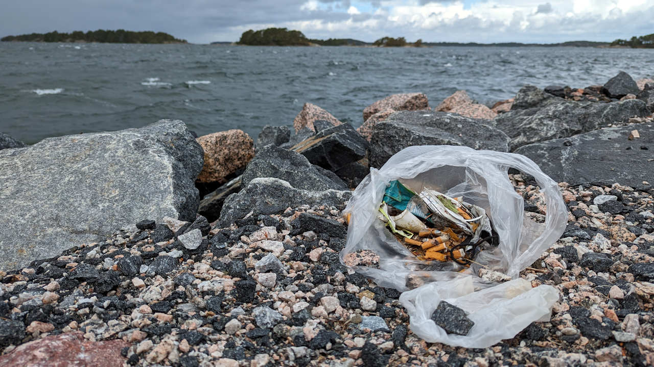 Eine weisse Tüte mit Zigarettenstumeln und anderen Abfällen an einem Kieselstrand. Im Hintergrund ist das graue Meer der Ostsee. 