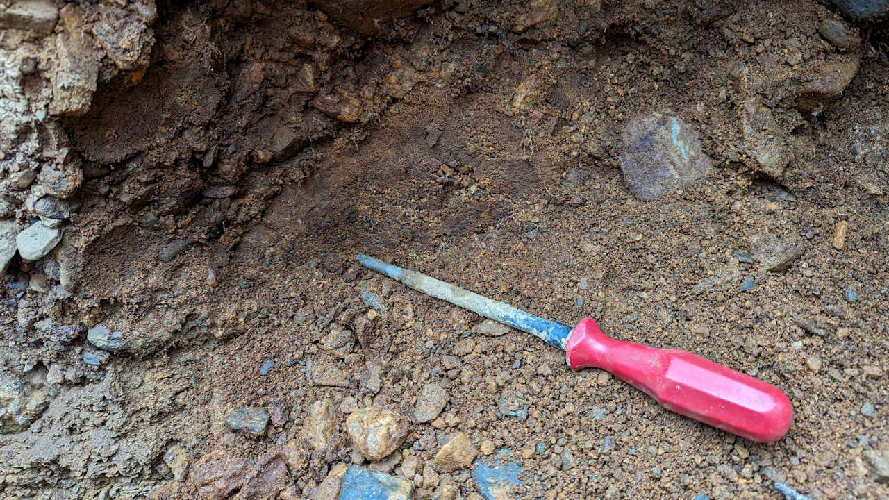 Ein Schraubenzieher mit rotem Griff liegt als Grabwerkzeug auf rostrotem goldhaltigen Kies Sand