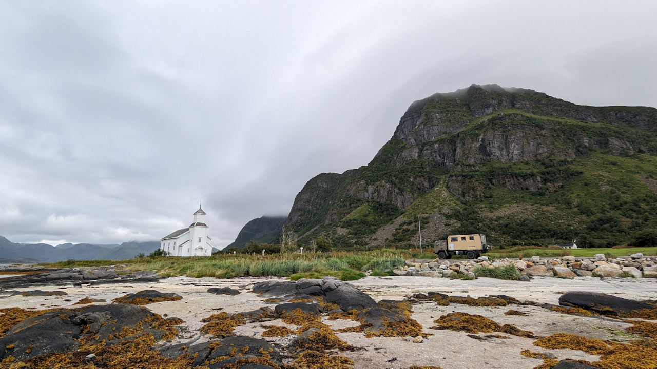 Panoramabild vom Grimsoysand, Lofoten Norwegen. Eine weisse Kapelle steht vor schroffen, wolkenverhangenen Bergen. 