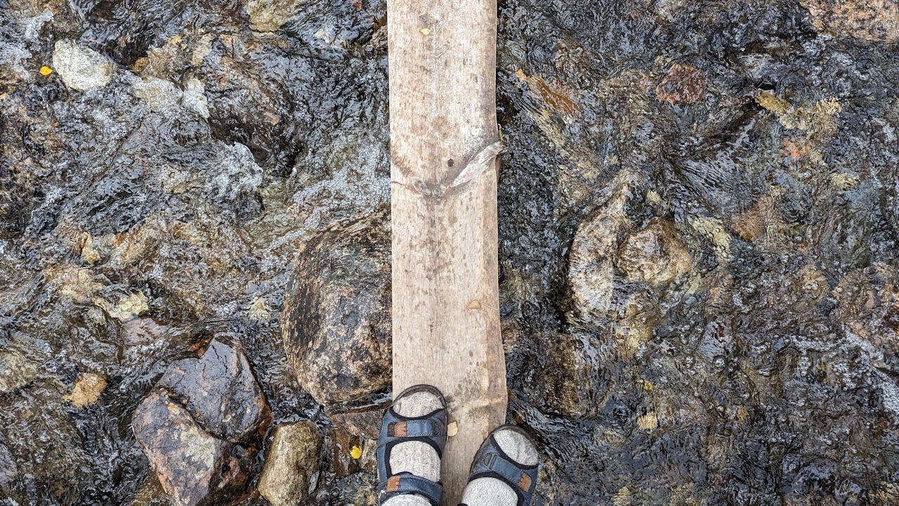 25 cm breiter Steg über einen kleinen Bach. Am unteren Ende sind zwei Füsse zu sehen.  