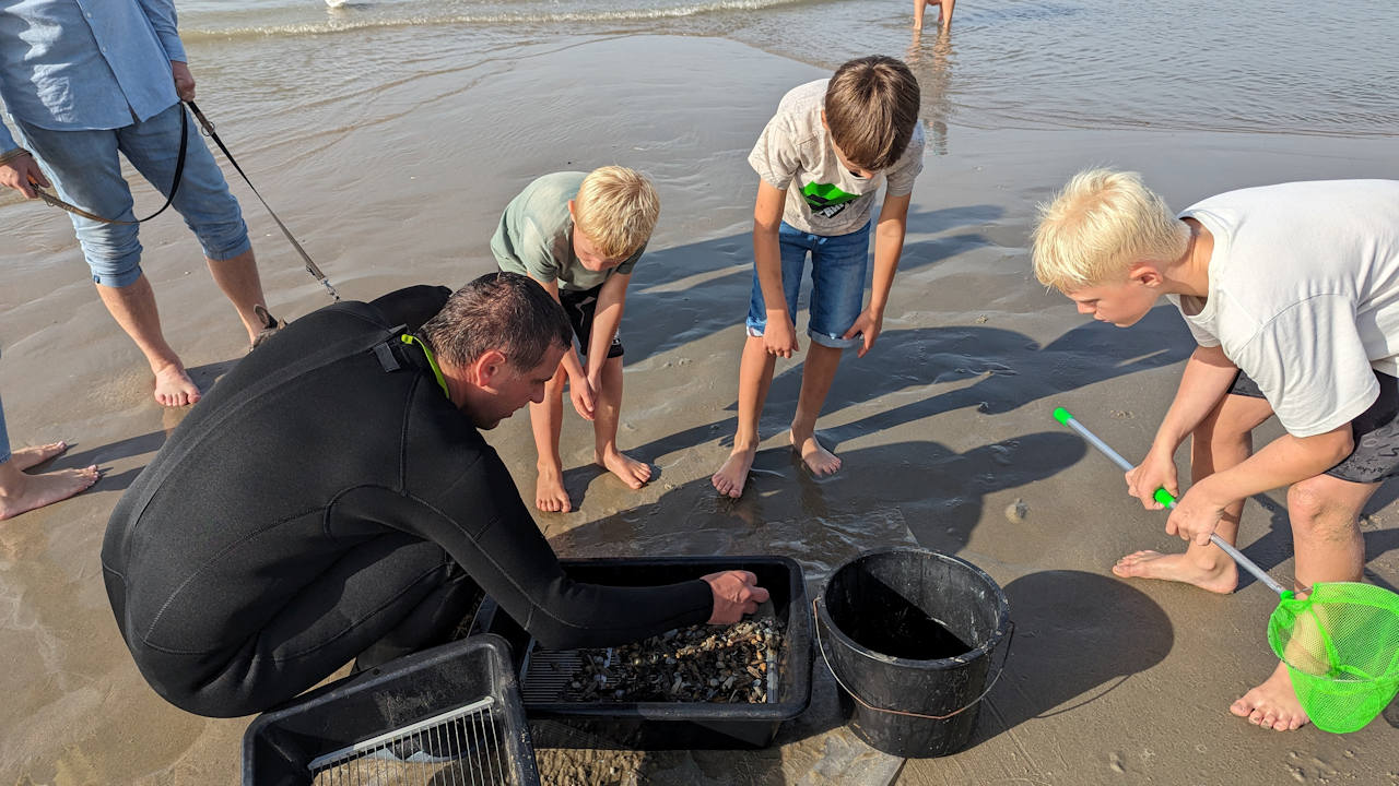 Ein Strandfischer sortiert in einer schwarzen Plastikwanne seinen Fang. umringt von drei neugierigen Kindern. 
