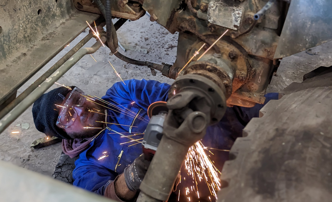 Ein Mechaniker im Blaumann und mit einer Schutzbrille verwendet eine Flex um eine Schraube an einer Antriebswelle aufzutrennen. Funken sprühen. 