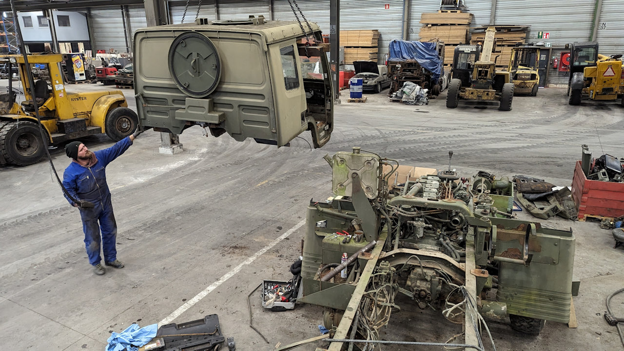 Ein grünes LKW Führerhaus schwebt in einer Halle mit drei Gabelstaplern und anderen gelagerten Sachen an einem Kran neben einem teilausgeschlachteten LKW