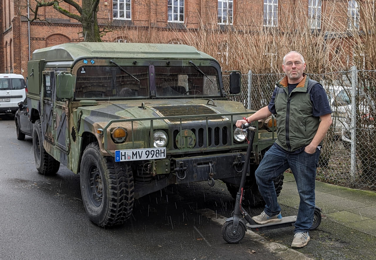 Ein Mann steht mit einem Elektro Scooter neben einem grünen US Army Humvee um diese miteinander zu vergleichen