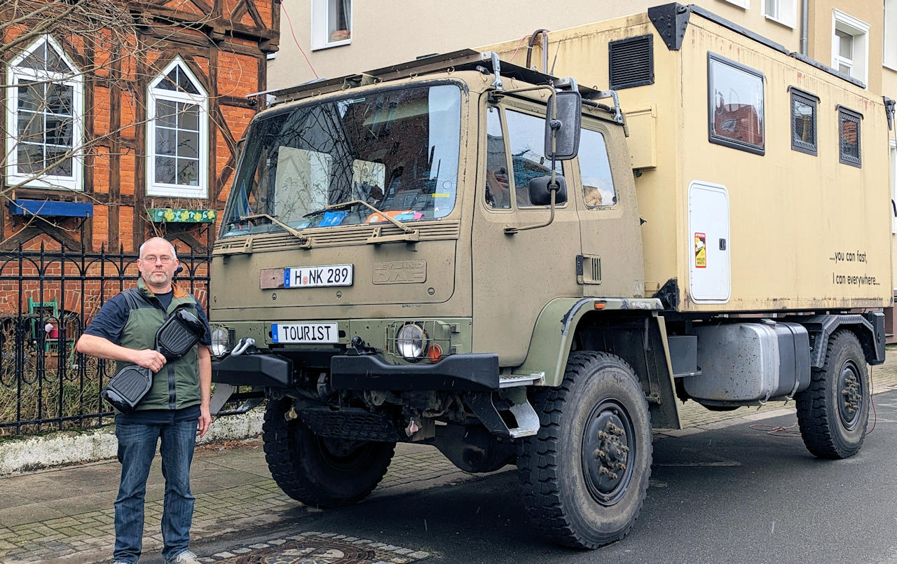 Ein Mann hält ein Elektro Hooverboard neben einem Allrad LKW in die Höhe um diese miteinander zu vergleichen