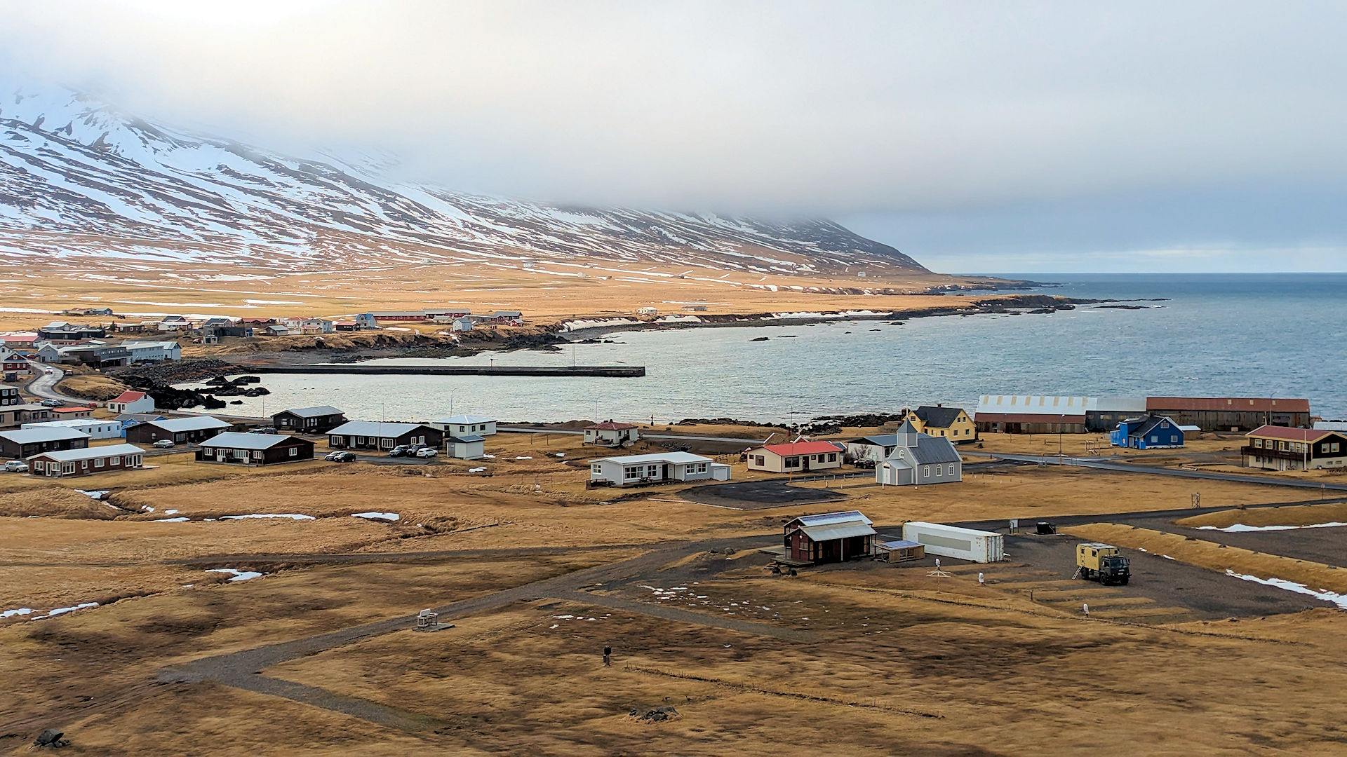 Der Ort Bakkagerdi mit Kirche, Hafen Pier und Campingplatz mit Meer und schneebedeckten Bergen im Hintergrund