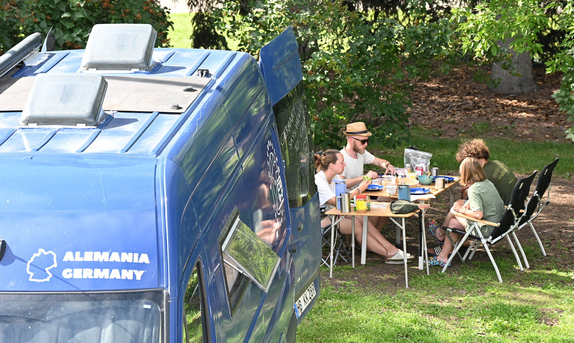 Gemeinsames Frühstück im Grünen. Vier deutsche Freunde neben einem Campingwagen auf dem Alemania Germany steht