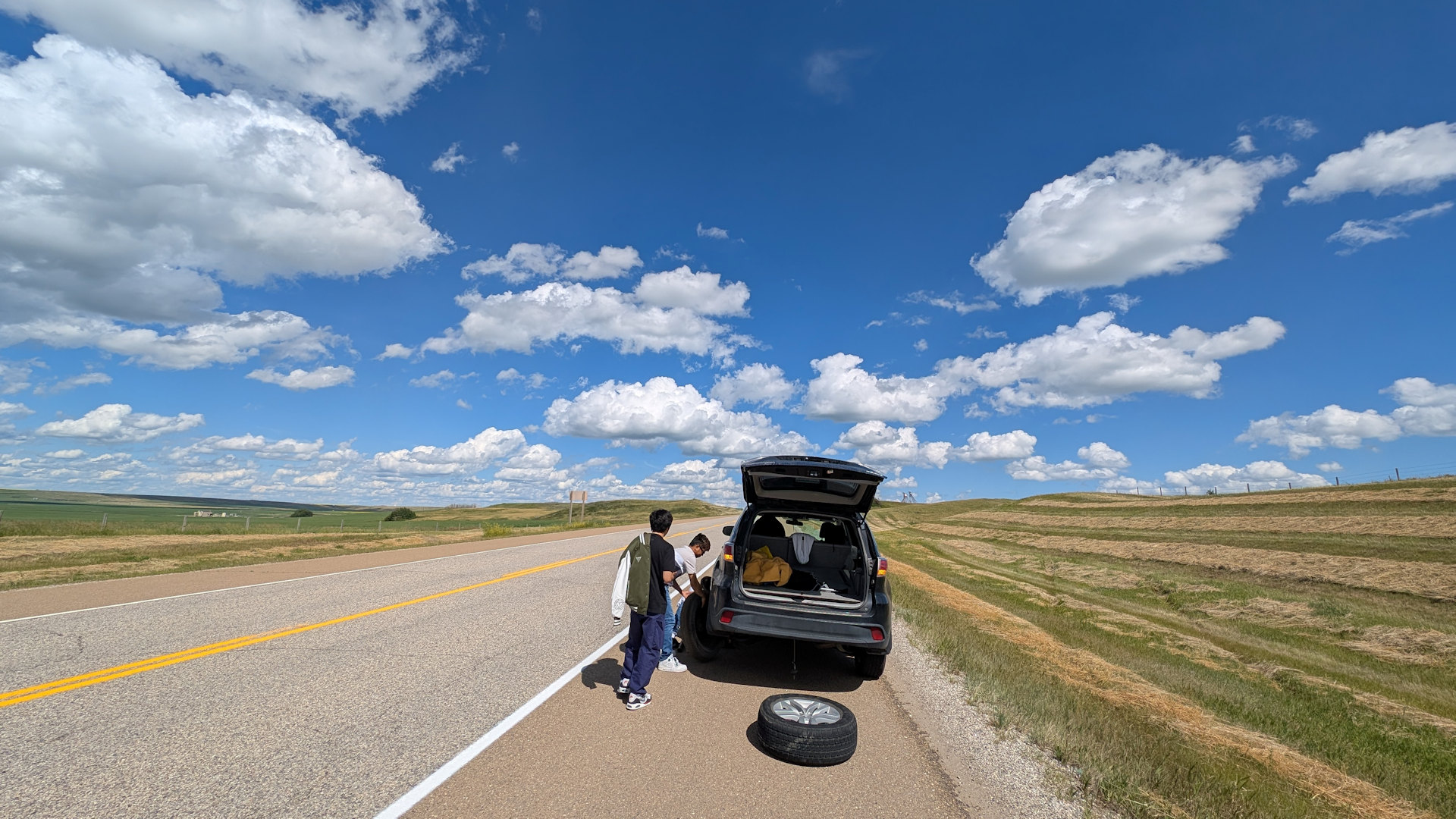 Drei Männer wechseln auf einer Landstrasse mit blauen Himmel und weissen Wolken ein Rad an einem Auto