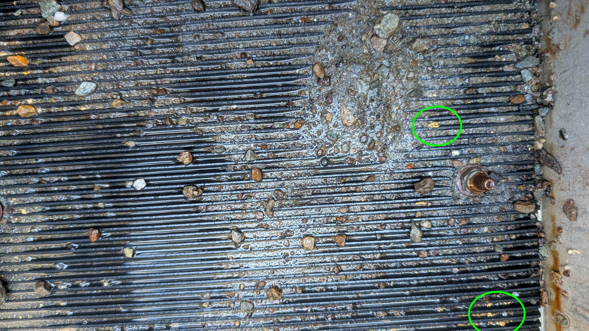 Schwarze Gummimatte mit Rillen einer Goldwaschrinne auf der kleine Steine und mit einem grünen Kreis markierte Goldnuggets liegen