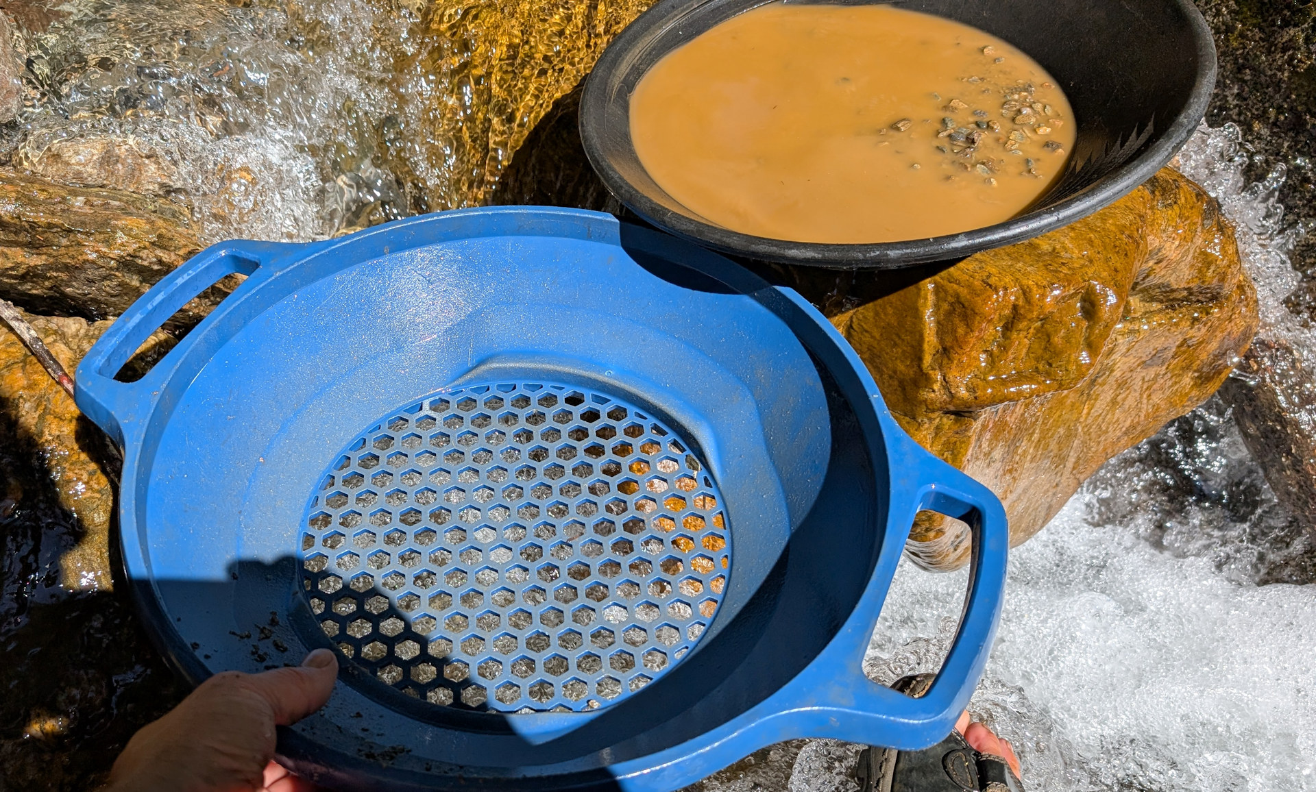 Blaues Sieb und schwarze Goldwaschpfanne mit braunem Wasser Sand Gemisch