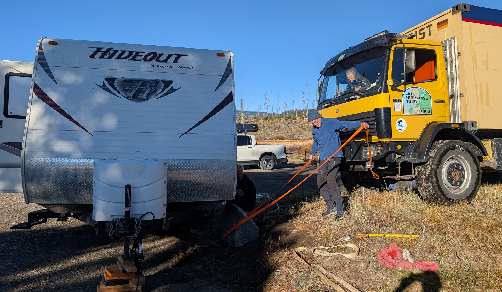 Ein orangener Spanngurt wird um einen Stein gespannt, der einen Campinganhänger blockiert 