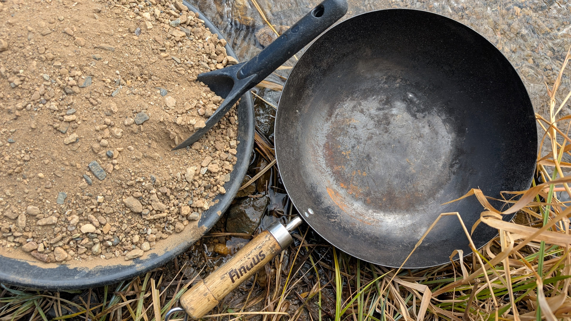 Goldwaschpfanne mit Goldsand neben einer grossen schwarzen Stahlpfanne mit Griff auf dem Findus steht