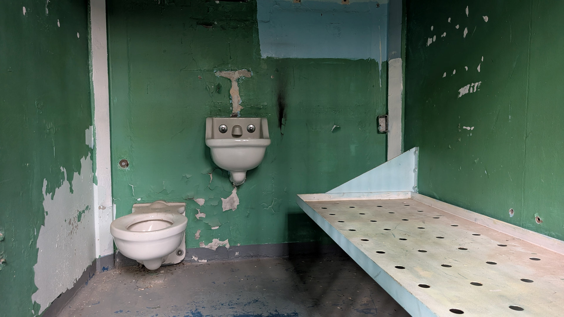 Gefängnis Zelle mit Pritsche, Klo, Waschbecken und grün gestrichener Wand 