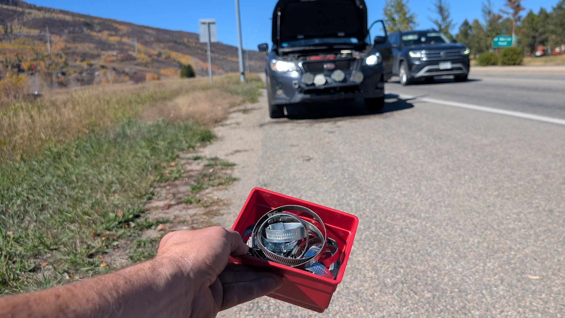 Eine Hand hält am Rande einer Schnellstrasse vor einem Auto mit geöffneter Motorhaube eine rote Box mit Schlauchschellen 