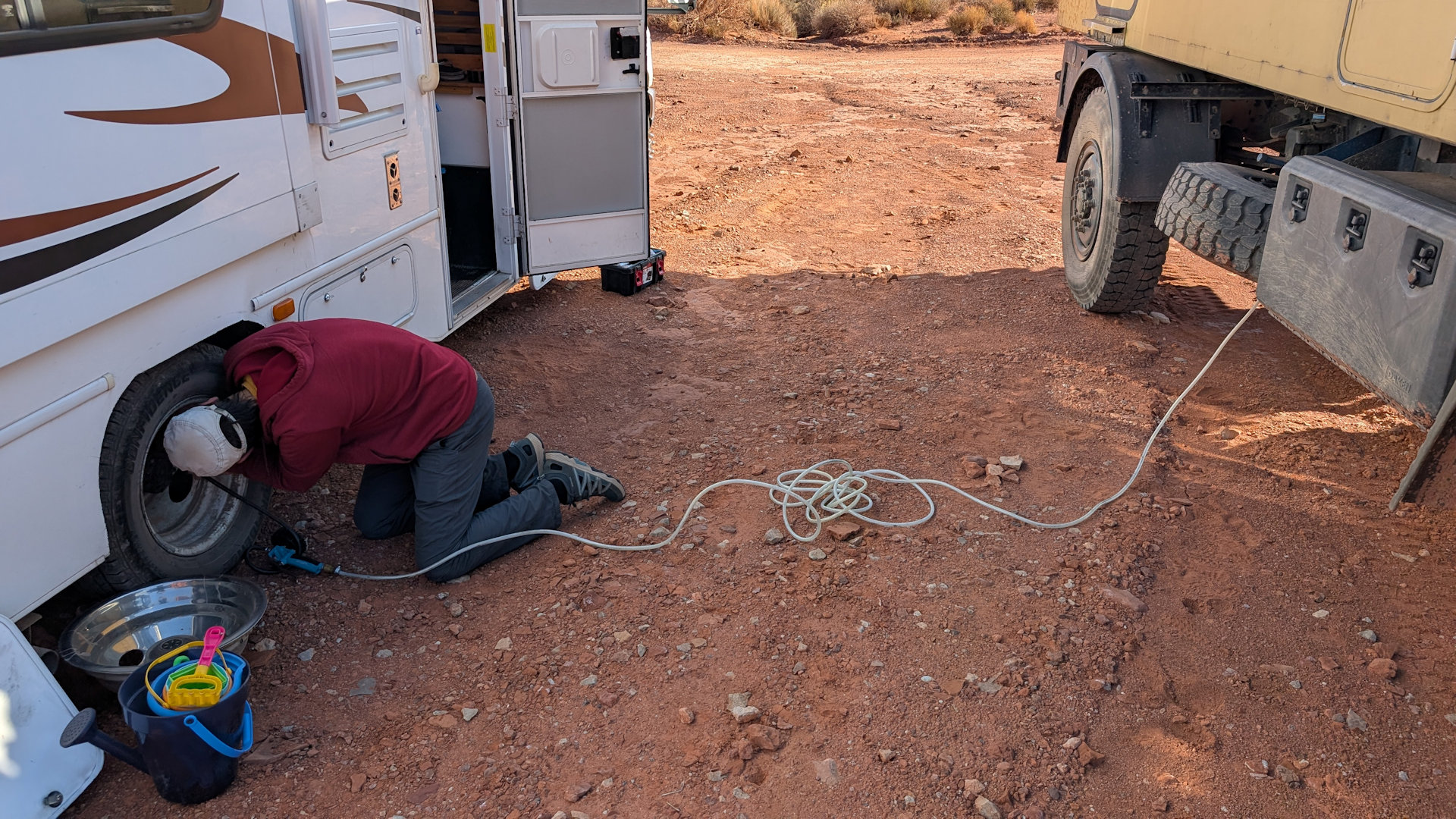 Ein Mann pumpt Reifen von einem weissen Camper mit Hilfe von einem Druckluftschlauch auf. Der Druckluftschlauch ist mit einem Offroad LKW verbunden. 