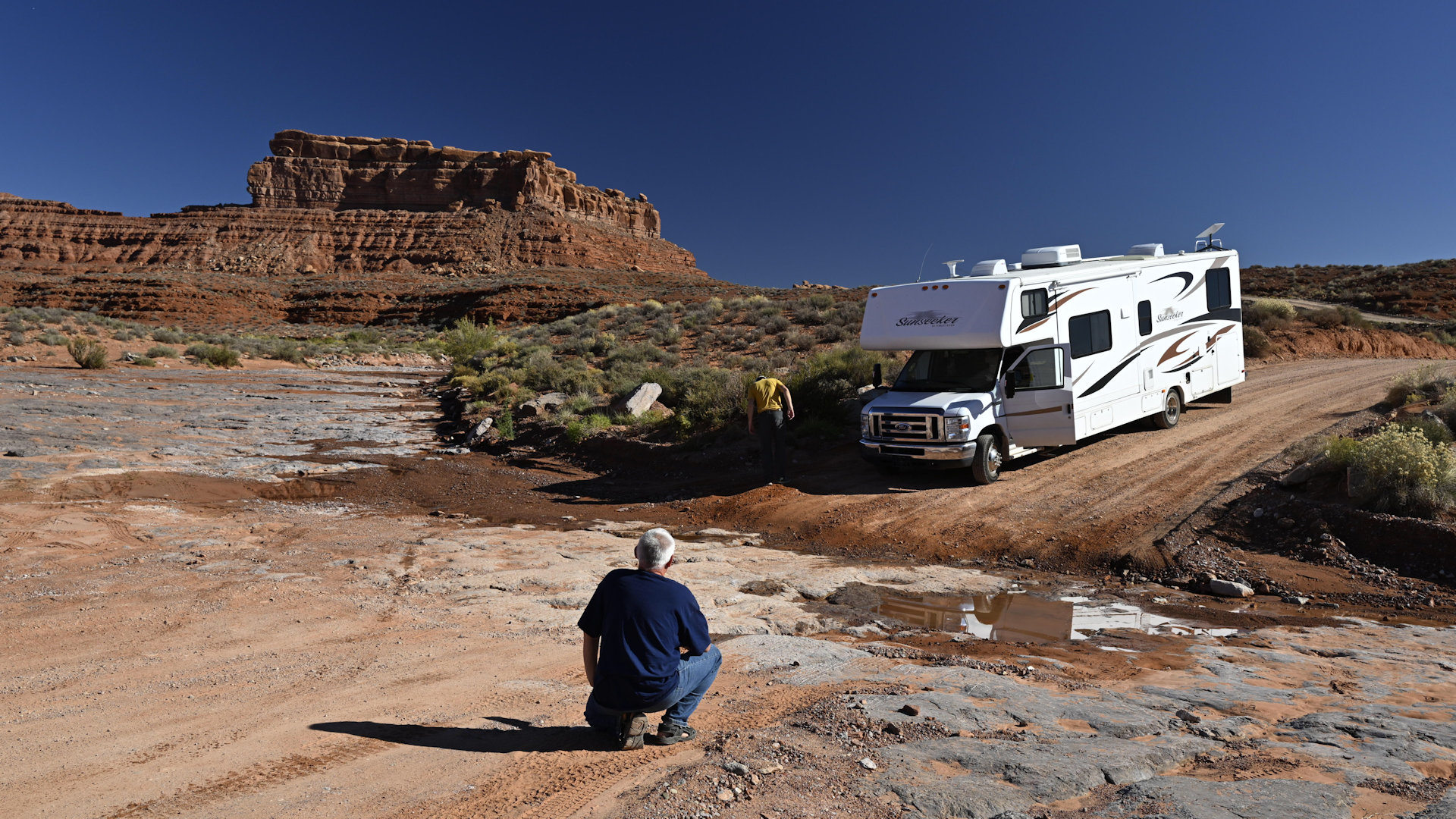 Ein weisser Camper fährt auf ein ausgetrocknetes Bachbett zu, vor dem ein Mann kniet. 