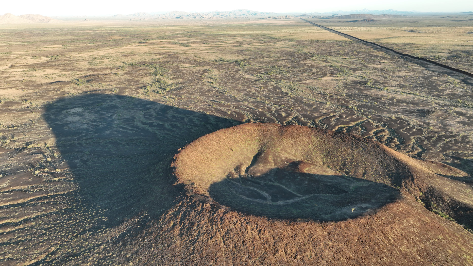 Vulkankegel in der Sonora Wüste mit langem Schatten und Grenz Zaun zu Mexiko im Hintergrund 