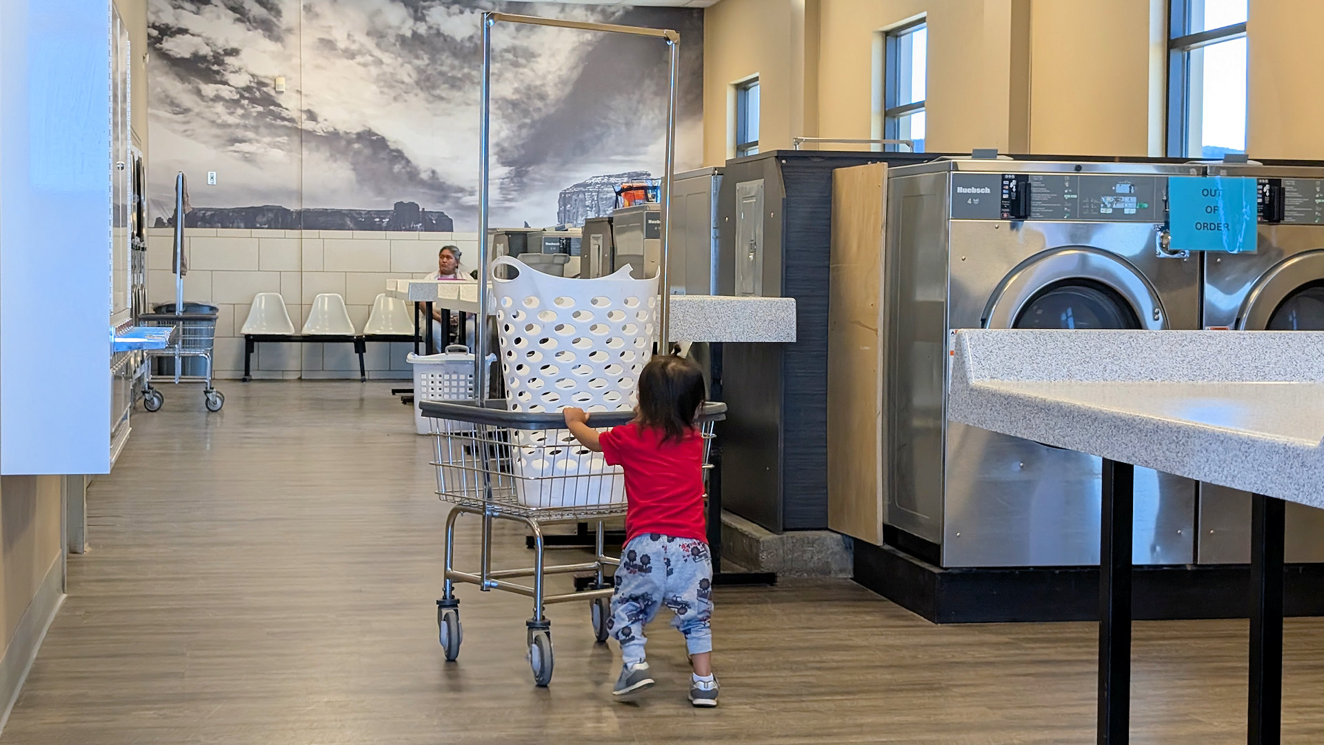 Kleinkind mit rotem Hemd schiebt einen rollbaren Wäschekorb durch einen Waschsalon.