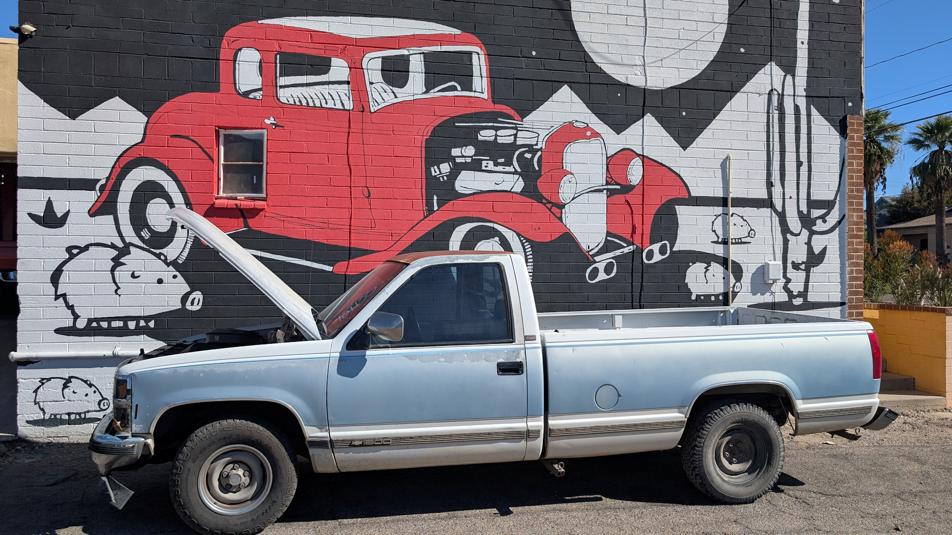 Eine Auto mit geöffneter Motorhaube parkt vor einem Wandbild mit roten Auto