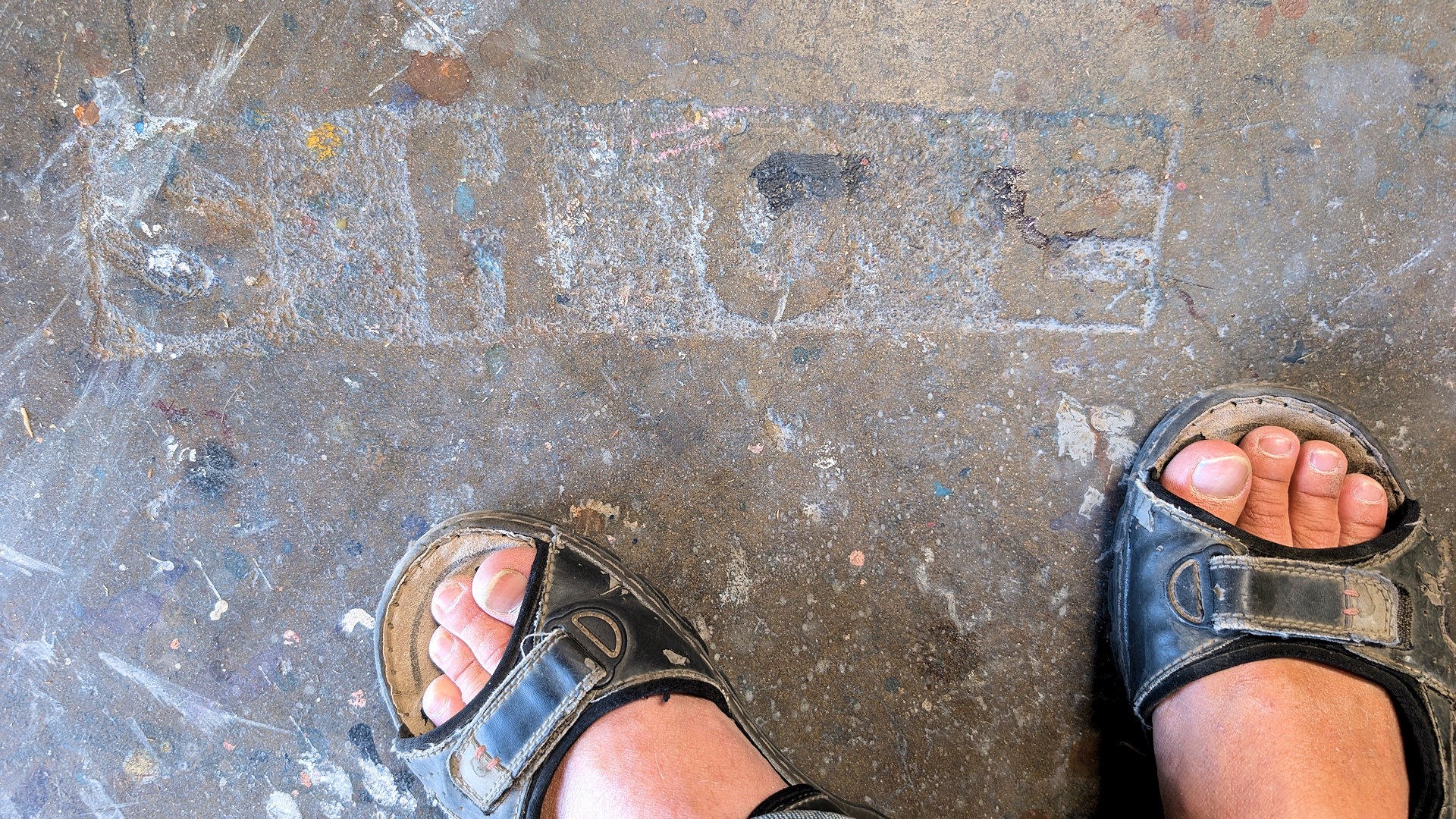 Auf einem Werkstattboden mit Farbkleksen sind die Buchstaben S H O E eingraviert. Daneben sind zwei Füsse in Sandalen zu sehen