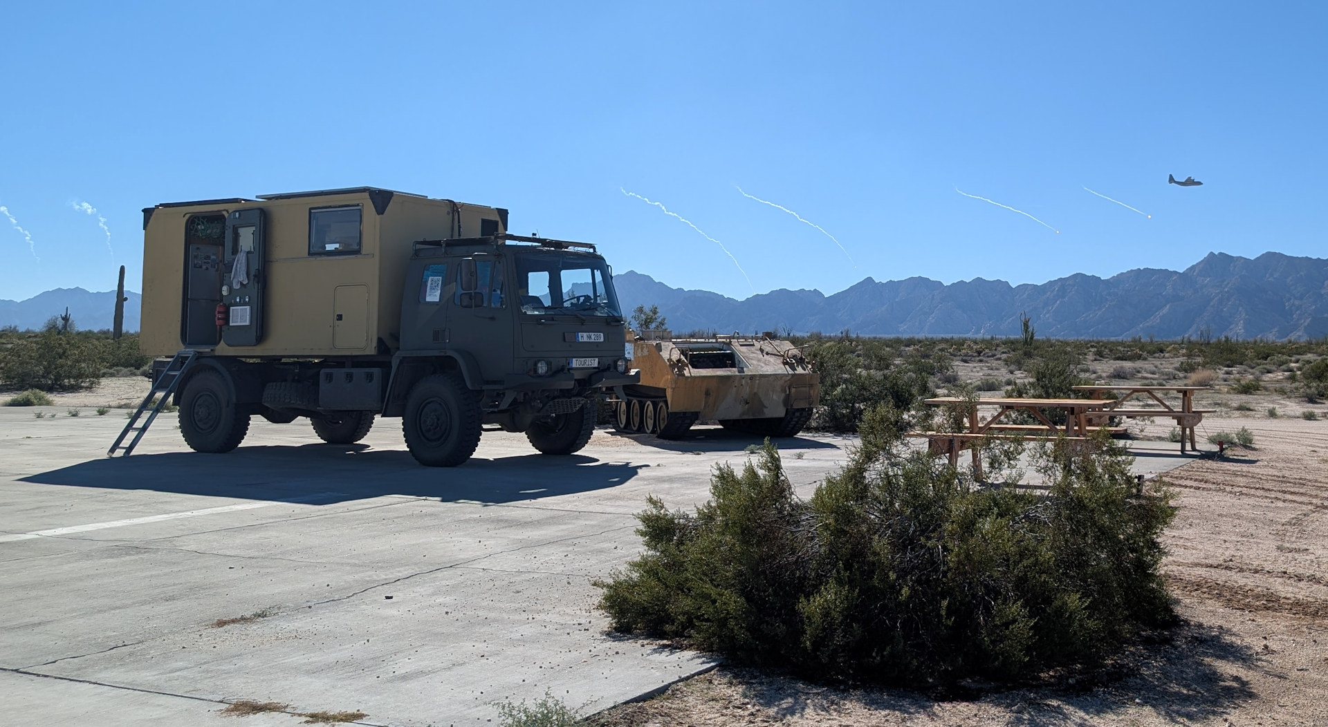Wüsten LKW parkt neben einem Panzerwrack während ein Transportflugzeug Flares abfeuert 
