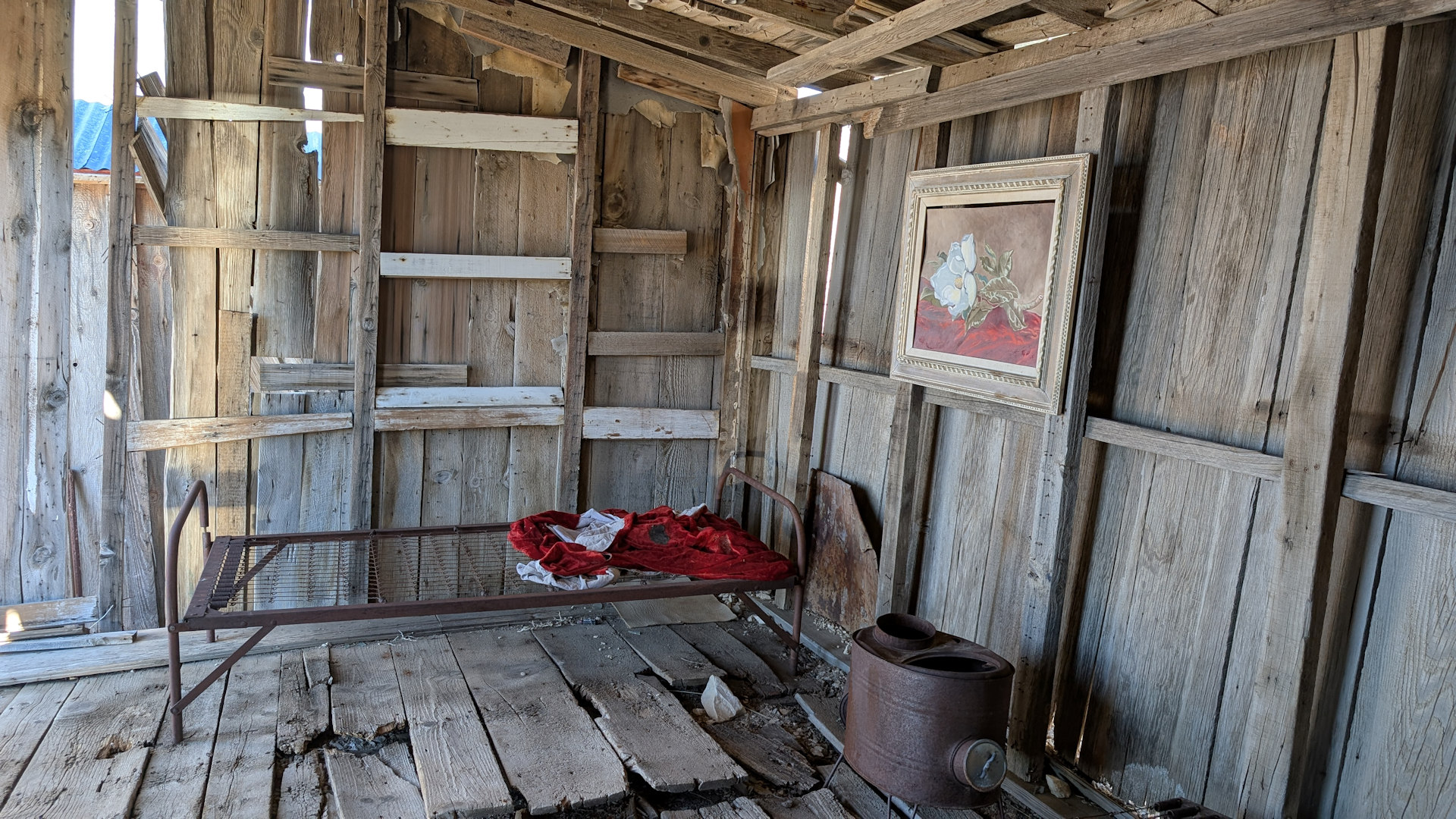 Im inneren einer Holzhütte in der ein rostiges Bett steht mit einem Bild an der Wand