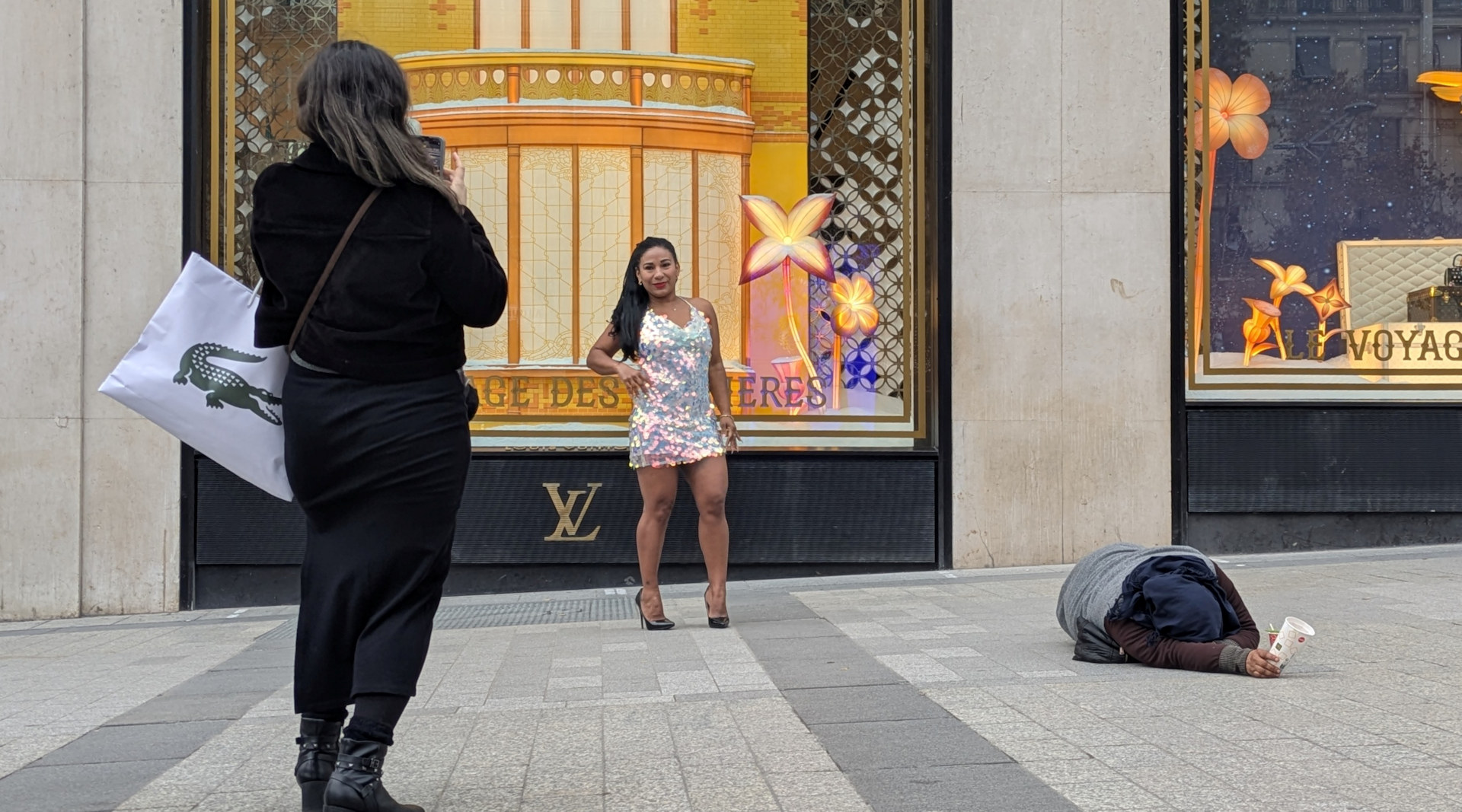 Frau mit einem schillernden Kleid wird in Paris vor einem Louis Vuitton Geschäft fotografiert während ein Bettler oder eine Bettlerin mit einem Pappbecher in der Hand neben ihr auf dem Boden liegt.