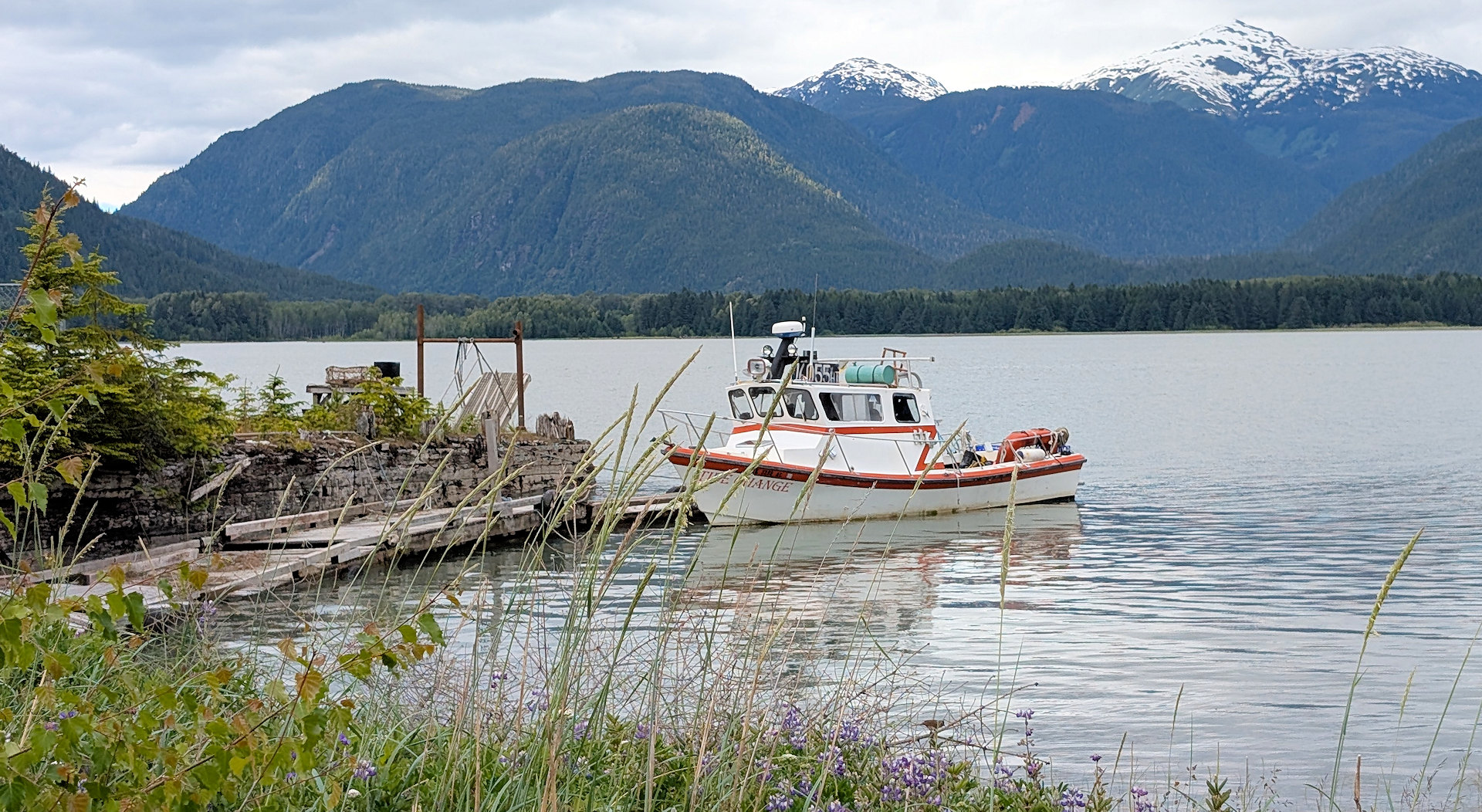 Das kleine Motorboot "High Tide" aus Anyox ist an einem Pier in Kitsault festgemacht. Schneebedeckte Berge sind im Hintergrund zu sehen.