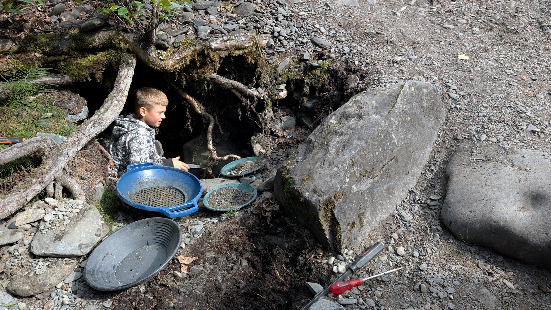 Neunjähriger Junge gräbt in einer kleinen Höhle nach Goldsand während mehrere Goldwaschpfannen und ein blaues Sieb davor liegen 