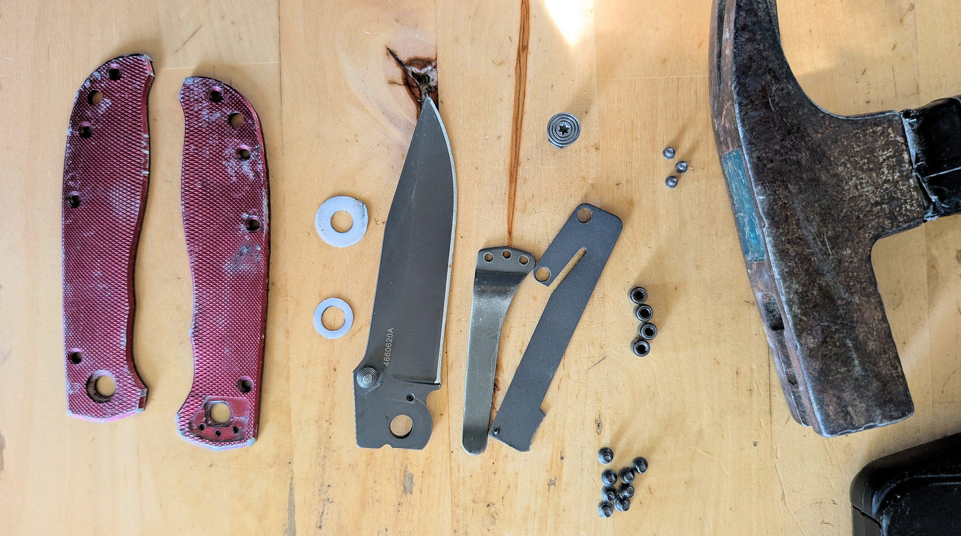 In Einzelteile zerlegtes Klappmesser mit Gehäuseteilen, Schrauben, Klinge und einem Hammer als Werkzeug daneben