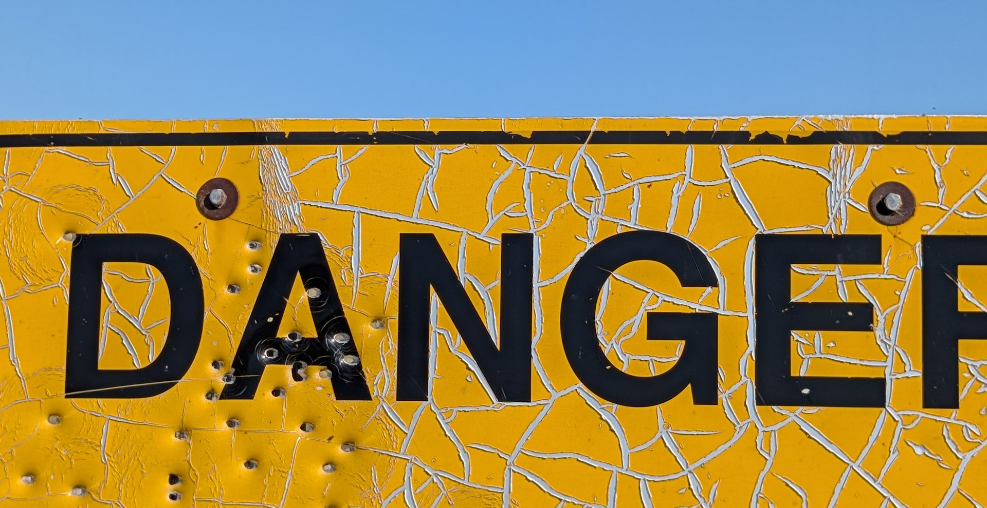 Gelbes Danger Schild bei dem die Farbe Risse bekommen hat und das durch Schrotkugeln getroffen wurde