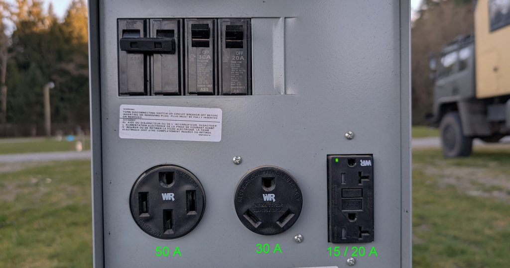 Stromsäule auf einem amerikanischen Campingplatz mit drei Steckdosen: links NEMA 14-50 (50 A), Mitte NEMA TT-30 (30 A), rechts NEMA 5-15 / 5-20 GFCI-Haushaltssteckdose (15/20 A), darüber die passenden Sicherungen, im Hintergrund der DAF T244.
