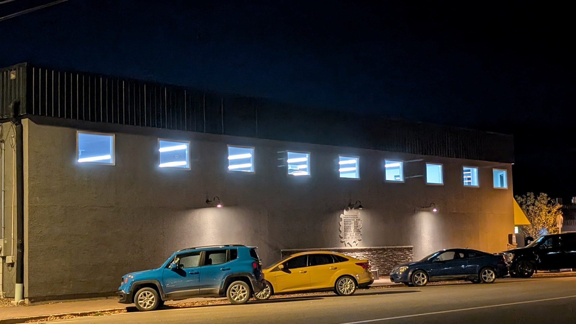Autos parken Nachts vor einer Halle bei der die Fenster mit Leuchtstoffröhren erleuchtet sind